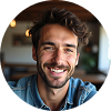 A smiling man with wavy brown hair and a beard, wearing a denim shirt, sits indoors with warm lighting in the background.