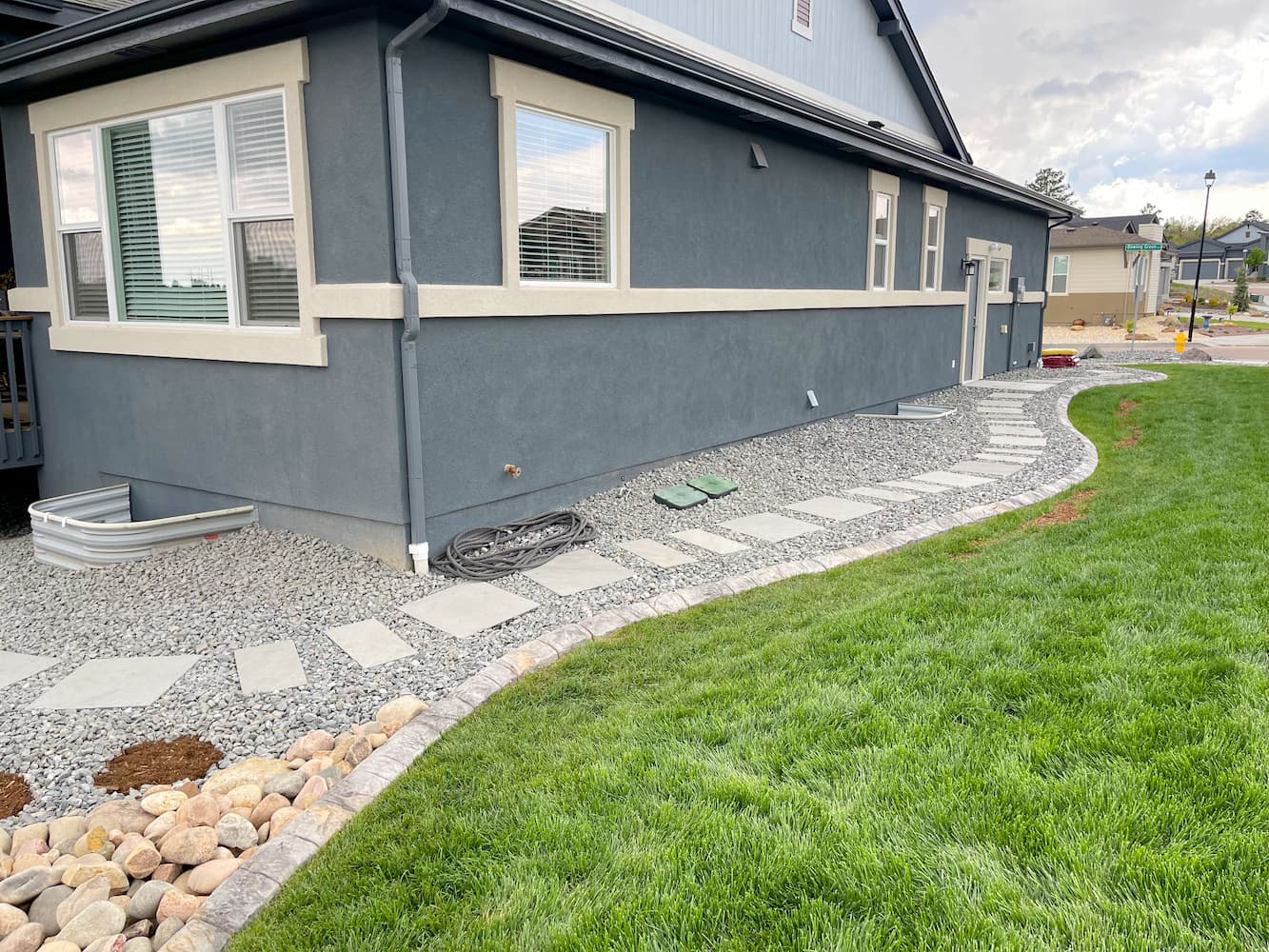 A modern house with dark blue siding features a stone walkway along its side, bordered by gravel and a green lawn. A small garden bed with rocks is visible near the walkway, and houses are in the background.