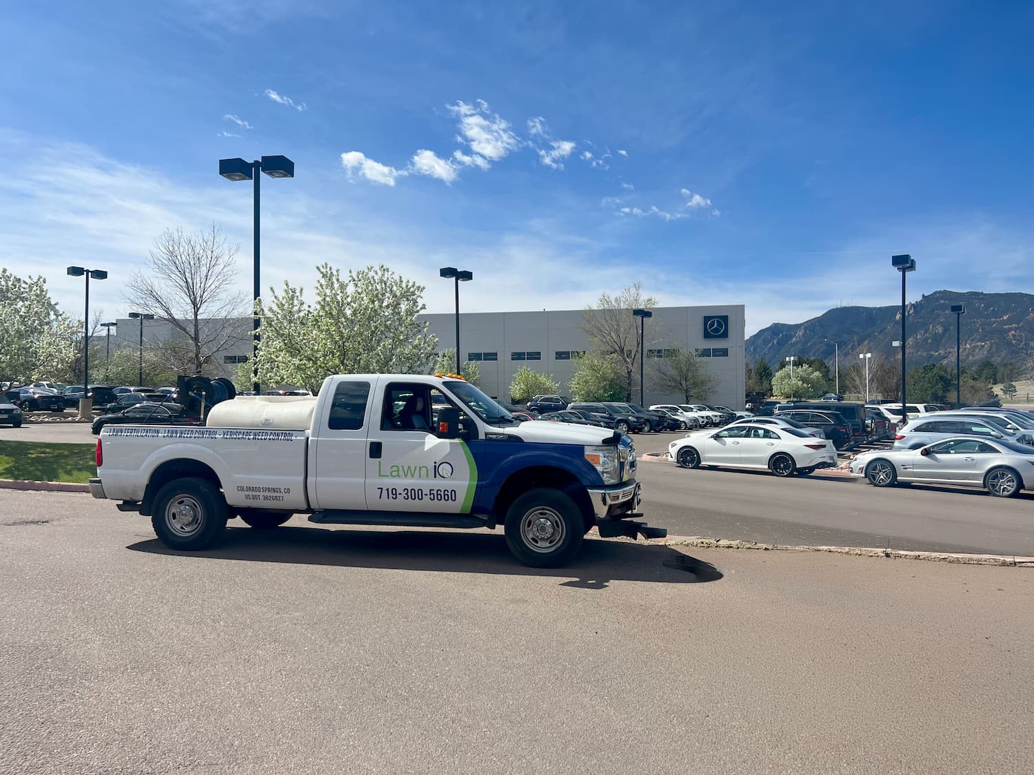 A white Lawn IQ pickup truck with lawn care equipment is parked on a street near a parking lot full of cars, with a large building and mountains in the background under a blue sky.