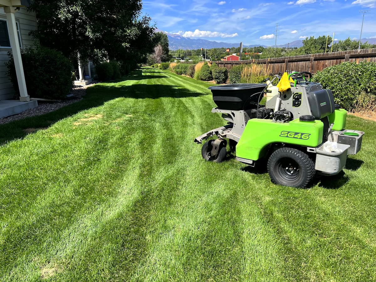 A green landscaping machine sits on a freshly mowed lawn in a sunny backyard, with striped grass patterns and trees lining the edges under a blue sky with scattered clouds.