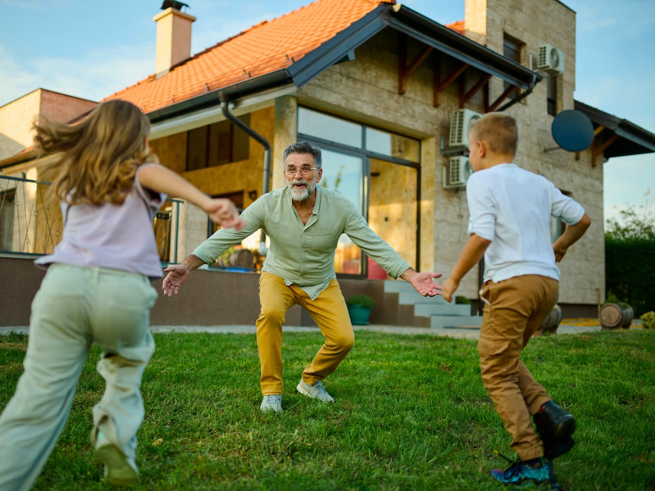 An older man with gray hair and a beard smiles with open arms as two children run toward him on the grass in front of a house. The scene is bright and cheerful, capturing a joyful family moment outdoors.