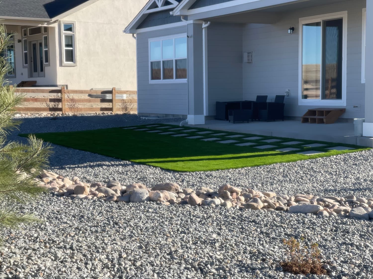 Modern house with gray siding, a covered front porch, black patio furniture, and a small patch of artificial grass bordered by square pavers. The yard is landscaped with rocks and a wooden fence in the background.