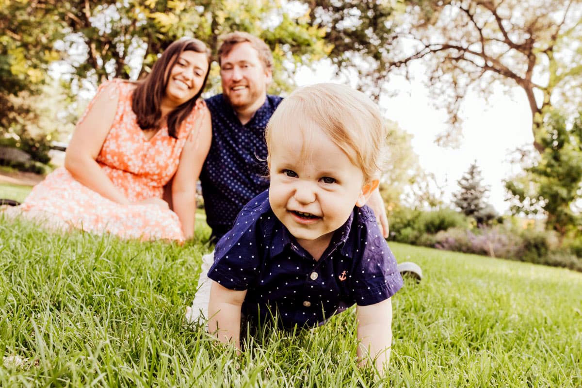 A smiling baby crawls on green grass in a park, with two smiling adults sitting in the background. Trees and bushes surround them on a sunny day.