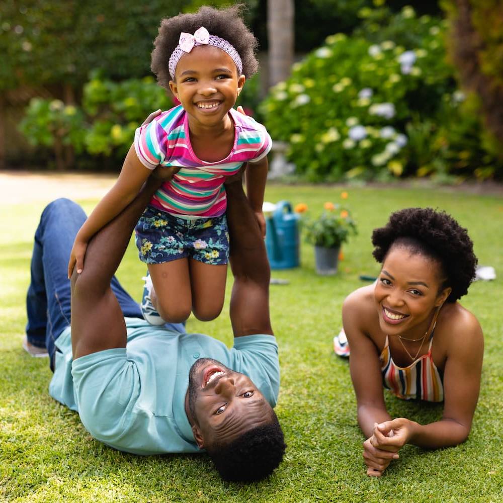 A smiling man lies on grass holding up a cheerful young girl, while a woman beside them smiles at the camera. They are outdoors in a garden with greenery and flowers in the background.