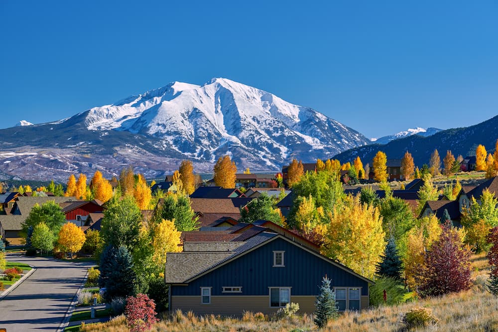 Colorful autumn trees surround houses in a residential neighborhood with a snow-capped mountain rising in the background under a clear blue sky.