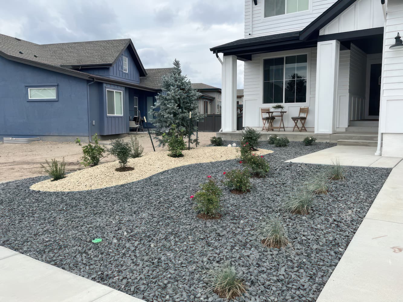 A front yard with xeriscape landscaping featuring gray gravel, beige rocks, small bushes, grasses, and a tree, in front of a white house with a porch and two wooden chairs. Another blue house is visible next door.